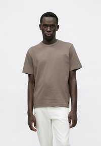 Καφέ κοντομάνικο μπλουζάκι με στρογγυλή λαιμόκοψη, φτιαγμένο από απαλό ύφασμα. Διαθέτει άνετη εφαρμογή και διακριτικές ραφές. Φορεμένο με ανοιχτόχρωμο παντελόνι.