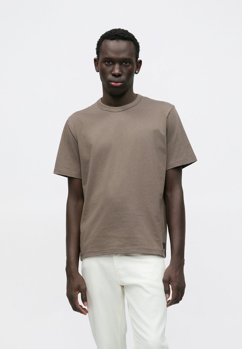 Καφέ κοντομάνικο μπλουζάκι με στρογγυλή λαιμόκοψη, φτιαγμένο από απαλό ύφασμα. Διαθέτει άνετη εφαρμογή και διακριτικές ραφές. Φορεμένο με ανοιχτόχρωμο παντελόνι.