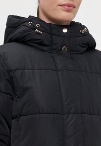 Svart puffjacka med hög krage, justerbar huva med guldfärgade detaljer, kviltad textur och tryckknappsstängning vid nacken.