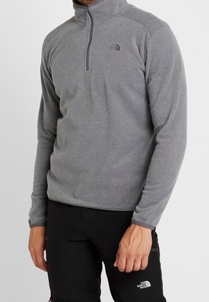 Homme portant un pull polaire gris à fermeture éclair quart et un pantalon noir, tous deux avec le logo The North Face, debout devant un fond uni.