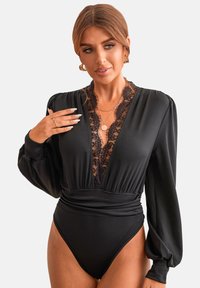 Černý body z jemného materiálu s krajkovými detaily na výstřihu a ramenou, s dlouhými rukávy, lichotivým střihem a vysokým vykrojením na nohách.