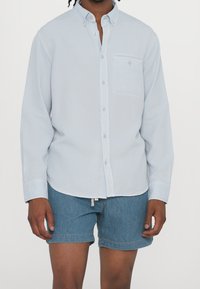 Světlomodrá košile s dlouhým rukávem, límcem na knoflíky, přední kapsou a zaobleným lemem, doplněná džínovými šortkami.