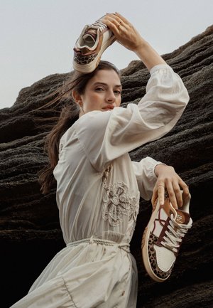 Las zapatillas presentan una base blanca con acentos de estampado de leopardo, detalles en marrón y rojo, y suelas de goma texturizadas, sostenidas por un modelo que lleva un vestido crema.