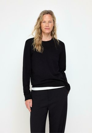 Lyshåret kvinde med bølget hår iført en sort langærmet sweater og sorte bukser, stående mod en ensfarvet hvid baggrund.