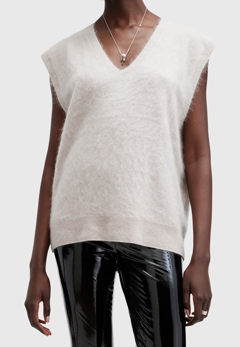 Personne à la peau foncée portant un gilet en laine blanc sans manches, un pantalon en vinyle noir brillant, un collier argenté et une bague à la main gauche.