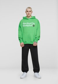 Sudadera verde con estampado de "New York City Basketball", bolsillo delantero, corte oversized, combinada con pantalones de chándal negros y zapatillas blancas.