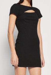 Femme portant une robe noire ajustée en maille côtelée, à manches courtes et avec une découpe horizontale sur la poitrine.