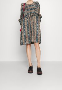 Šaty s květinovým vzorem v tmavě modrozelené barvě s volánovými rukávy a nařaseným pasem. Kombinace s černou obuví. Pozadí je jednoduché bílé.