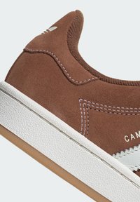 Brown suede sneaker with white rubber sole, featuring textured upper, contrasting white stitching, and a logo detail near the heel.