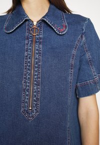 Denimklänning med en framdragkedja, spetsig krage och korta ärmar. Kontraststygn i röd färg med en dekorativ detalj på kragen.