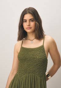 Vihreä smokkihihainen mekko ohuilla hihnoilla, teksturoidulla yläosalla ja liehuvalla helmalla. Mekossa on kiiltäviä yksityiskohtia kaula-aukossa ja ranteessa.