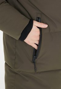 Olivgrön vadderad jacka med en sidoficka med dragkedja, ribbade svarta muddar och slät tygstruktur. Närbild av hand i fickan.