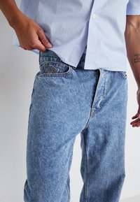 Jeans en denim bleu clair avec une texture légèrement délavée, dotés d'un zip à boutons et d'un logo brodé sur la poche arrière, associés à une chemise rayée.