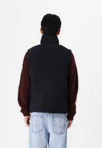 Zwarte corduroy gilet met hoge kraag, gedragen over een bruine gestructureerde overhemd met lange mouwen. Lichtblauwe jeans met eenvoudige steken op de achterzak.