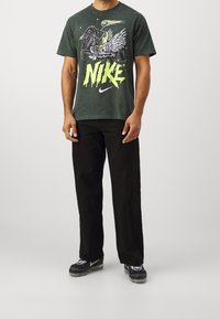 Camiseta gráfica verde con un diseño de caballo y alas, que presenta el texto amarillo "NIKE". Pantalones negros con detalles texturizados y calzado gris.