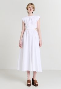 Donna che indossa un vestito midi bianco senza maniche con vita arricciata e ritagli, abbinato a sandali con zeppa marroni, in piedi su uno sfondo chiaro e uniforme.