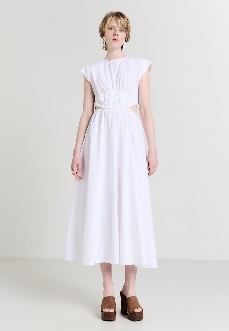 Donna che indossa un vestito midi bianco senza maniche con vita arricciata e ritagli, abbinato a sandali con zeppa marroni, in piedi su uno sfondo chiaro e uniforme.