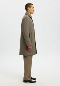 Cappotto in misto lana con motivo houndstooth beige chiaro, caratterizzato da un alto colletto, bottoni e tasche laterali, abbinato a pantaloni beige e scarpe nere.