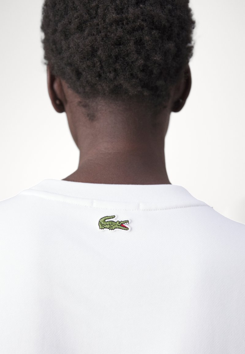 Camisa polo blanca con cuello plano, que presenta un pequeño logo de cocodrilo bordado en verde en la parte posterior del cuello. Textura de cabello corto visible.