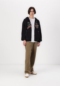 Svart oversized hettegenser med rødt og hvitt grafisk design, kombinert med en hvit t-skjorte og løse khakibukser, iført hvite sneakers.