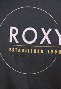 Juodas medvilninis marškinėliai su „ROXY“ baltu tekstu ir „ESTABLISHED 1990“ auksiniu, aplink tekstą yra apvalus baltas bruožas.