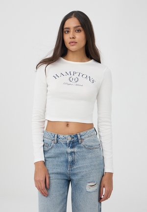 Biały krótki top z długim rękawem z granatowym nadrukiem "HAMPTON'S Perfect Match", zestawiony z wysokim stanem, zniszczonymi jasnymi dżinsami.