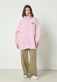 Roosa, karvane fliispusa kõrge kraega, esise paigaldatava lukuga ja värvilise plaastriga; kombineeritud beeži laia säärega pükste ja valgete tossudega.