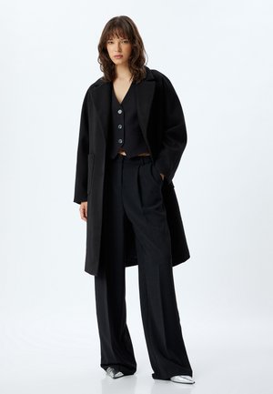 Sort oversized frakke, lagdelt over en sort vest med knapper, parret med skræddersyede sorte bukser og sølvfarvede sko. Glat stof og struktureret pasform.