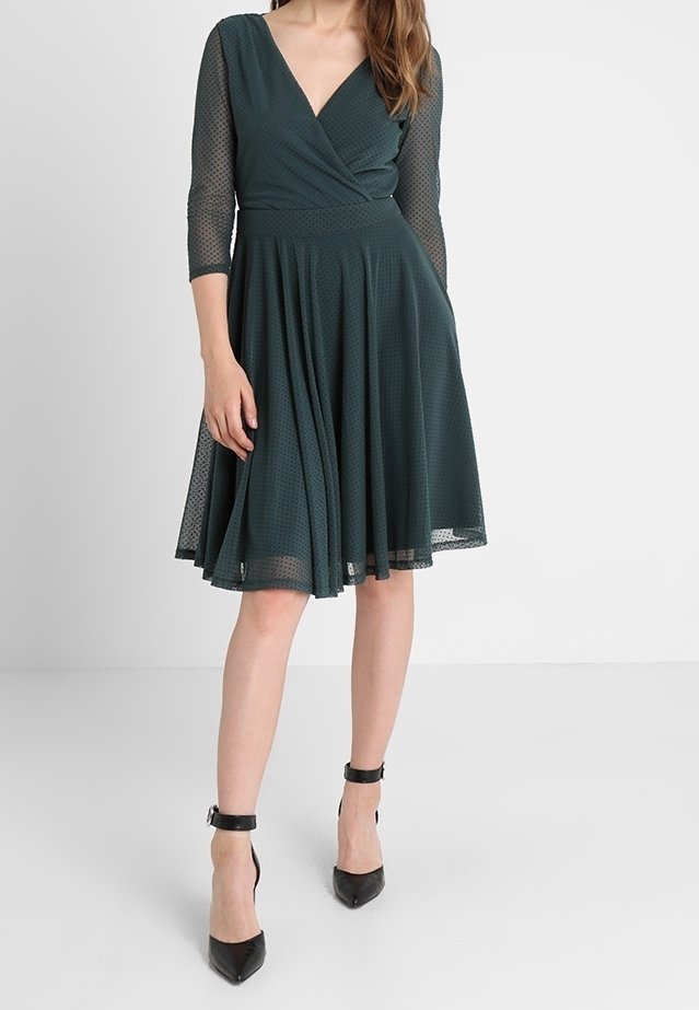 Femme portant une robe vert foncé longueur genou avec des manches trois-quarts en tulle et des escarpins noirs à bride cheville.