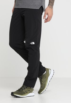 Man draagt zwarte North Face broek en olijfgroene Salomon sneakers, staand tegen een effen lichte achtergrond.