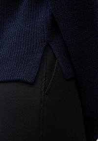Gros plan de l'ourlet d'un pull en maille bleu marine avec une fente latérale au-dessus de la poche d'un pantalon noir.