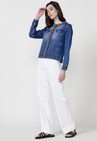 Jeans jakna s okruglim izrezom, prednjim džepovima i teksturiranim rubom. Model nosi bijele hlače širokih nogavica i crne sandale.