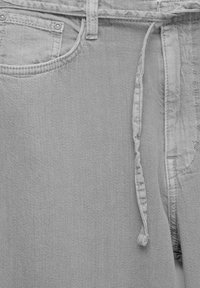 Gros plan sur un pantalon en denim gris clair avec une poche avant, une passante de ceinture et un cordon en tissu à la taille.