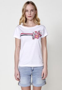 Camiseta blanca de manga corta con estampado floral en rosa y verde, acompañada de rayas rojas y navy cerca del escote. Tejido de algodón suave.