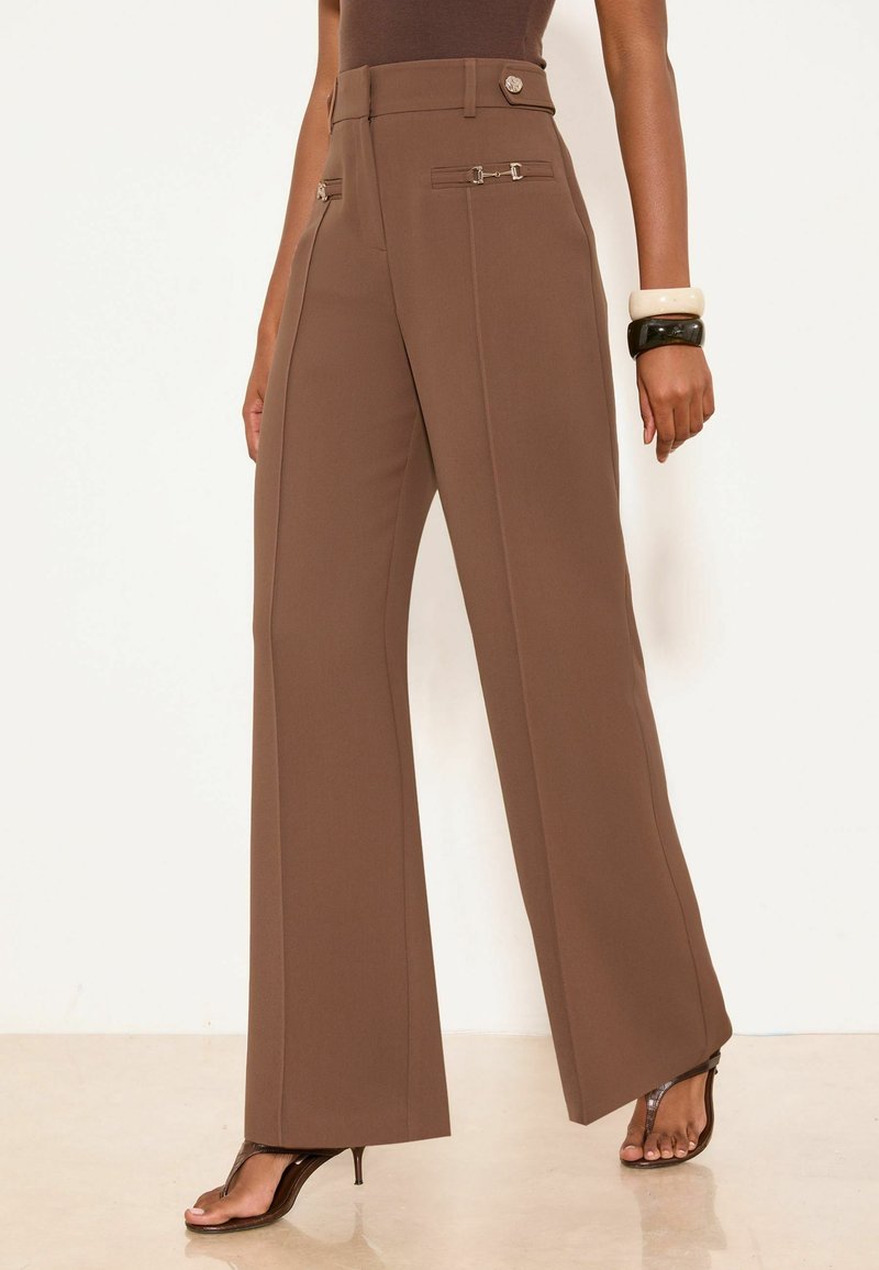 Brown high-waist wide-leg trousers with front decorative fastenings, worn with open-toe heels and bracelets on the right wrist.
