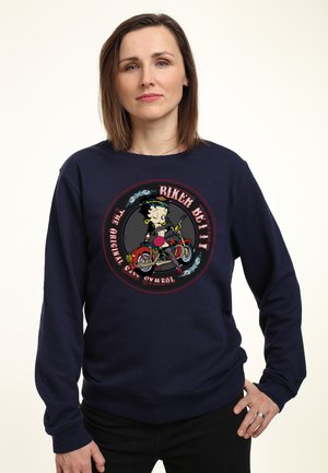 Kvinde med lige brunt hår iført en navy sweatshirt med en biker-tema Betty Boop grafik og tekst på fronten.