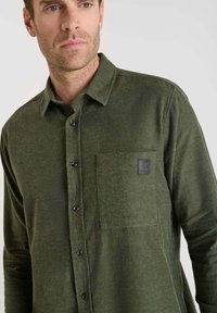 Man in een donkergroen overhemd met knopen, voorzien van een borstzak en een klein zwart logo-opdruk op de zak.
