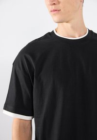 Tricou din bumbac negru, cu tiv alb în jurul gâtului și pe mânecile scurte. Croi lejer cu o textură netedă.