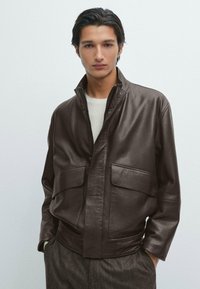 Giacca di pelle marrone con chiusura a zip, colletto alto e due grandi tasche frontali. Texture morbida, vestibilità rilassata, abbinata a un maglione leggero.