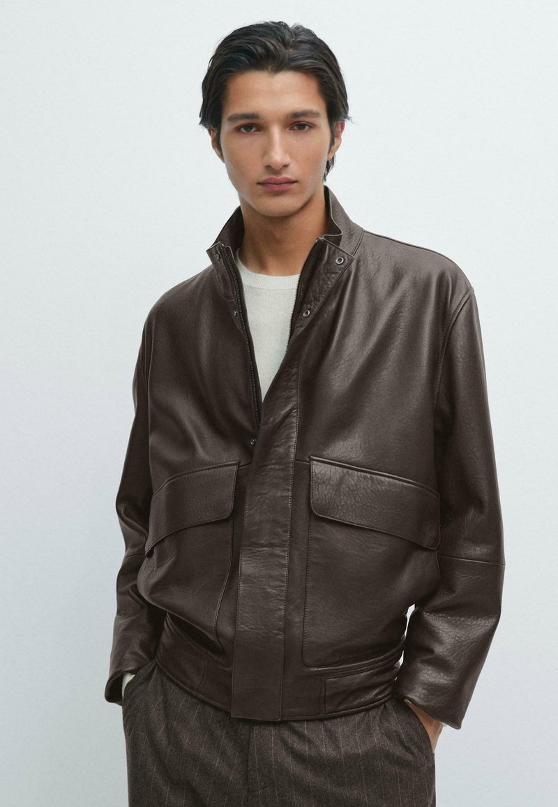 Giacca di pelle marrone con chiusura a zip, colletto alto e due grandi tasche frontali. Texture morbida, vestibilità rilassata, abbinata a un maglione leggero.