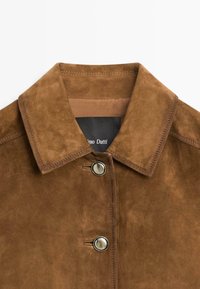 Colletto di una giacca in camoscio marrone con due bottoni a pressione in metallo e cuciture visibili, che mostrano parte dell'etichetta interna.