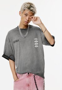 Ung person med kort, bleget hår iført en falmet grå oversized t-shirt med trykte læber og "TRUE SELF", lyserøde jeans, sølvkæder, der rører ved øjet.