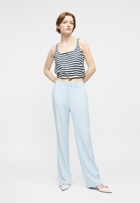 Γυναίκα που στέκεται με τα χέρια πίσω από την πλάτη, φοράει ριγέ αμάνικη μπλούζα, ανοιχτό μπλε παντελόνι με φαρδιά πόδια και λευκά μυτερά παπούτσια.