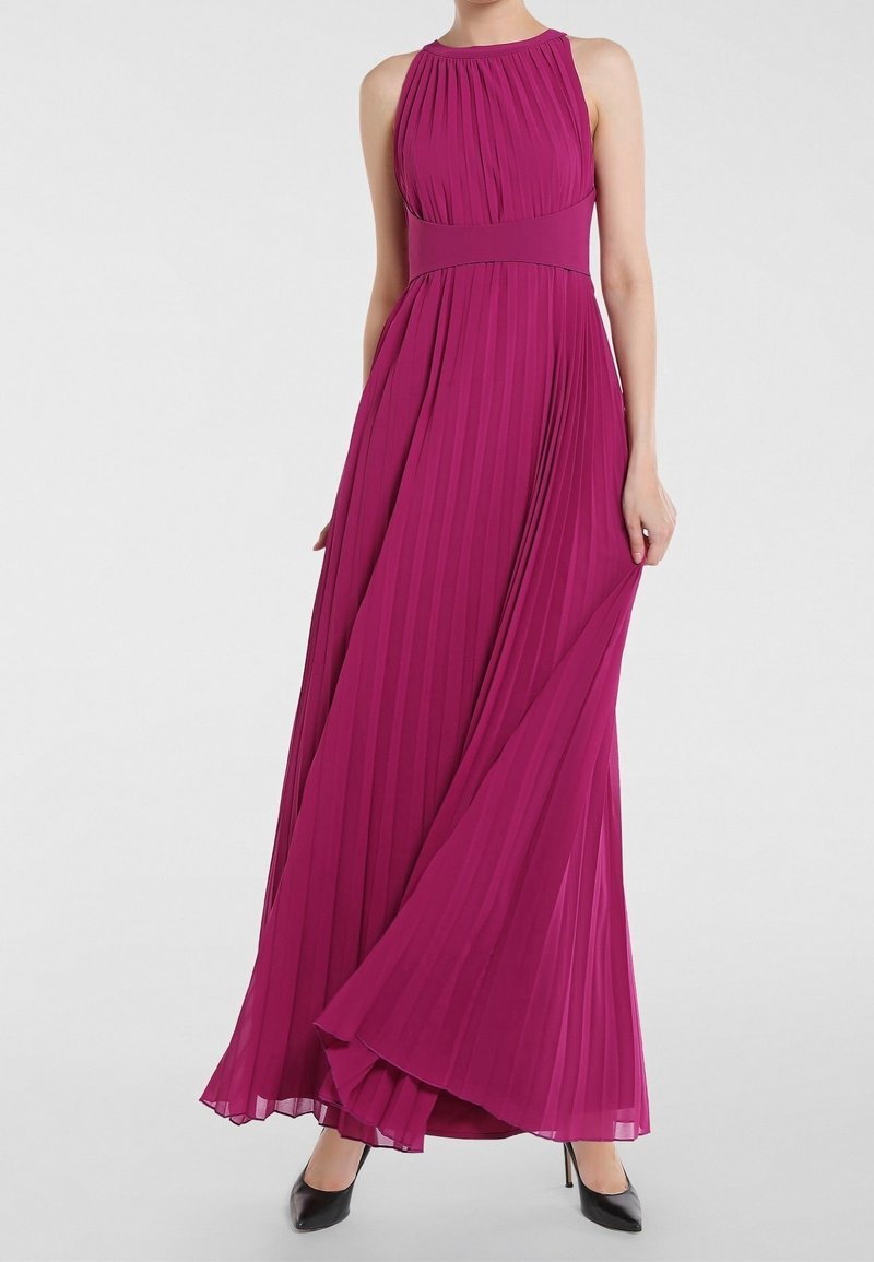 Robe longue plissée magenta avec un col noué autour du cou, une taille cintrée, une jupe fluide et une texture lisse ; associée à des chaussures à talons hauts noires.