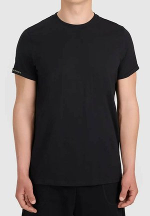 Mężczyzna w jednolitej czarnej koszulce z krótkim rękawem z podwiniętymi rękawami, stojący na neutralnym tle.