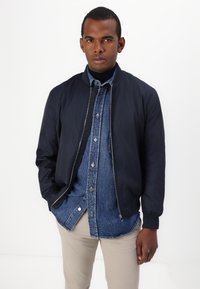 Mørkeblå bomberjakke med glidelås, brukt over en lys blå denimskjorte og beige bukser. Har ribbestrikkede mansjetter og krage.