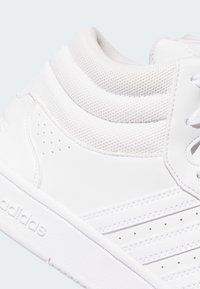 Detailný záber bielych vysokých tenisiek Adidas so polstrovaným golierom okolo členku, perforovanými koženými prúžkami a vyrazeným logom na podrážke.