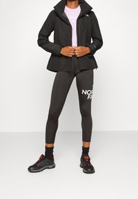 Persoon in een zwarte buitenjas, zwarte legging met wit logo, zwarte sokken en zwarte wandelschoenen met rode accenten.