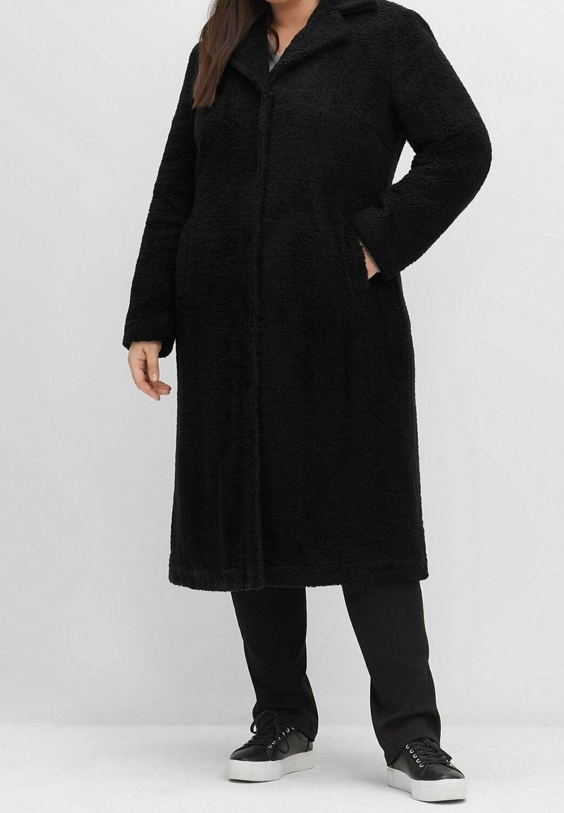 Musta pitkä takki, joka on valmistettu pehmeästä, teksturoidusta materiaalista, jossa on kaulus, etunapit ja sivutaskut. Käytetty mustien housujen ja tennareiden päällä.