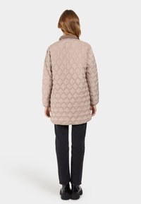 Lichtbeige gewatteerde jas met een gestructureerde afwerking, voorzien van een kraag en lange mouwen, gedragen over zwarte broek en zwarte laarzen.